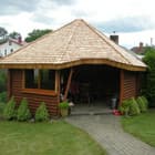 Rąstinė altana su šviežiomis kedro čerpėmis – priešakyje sodas, fone namas