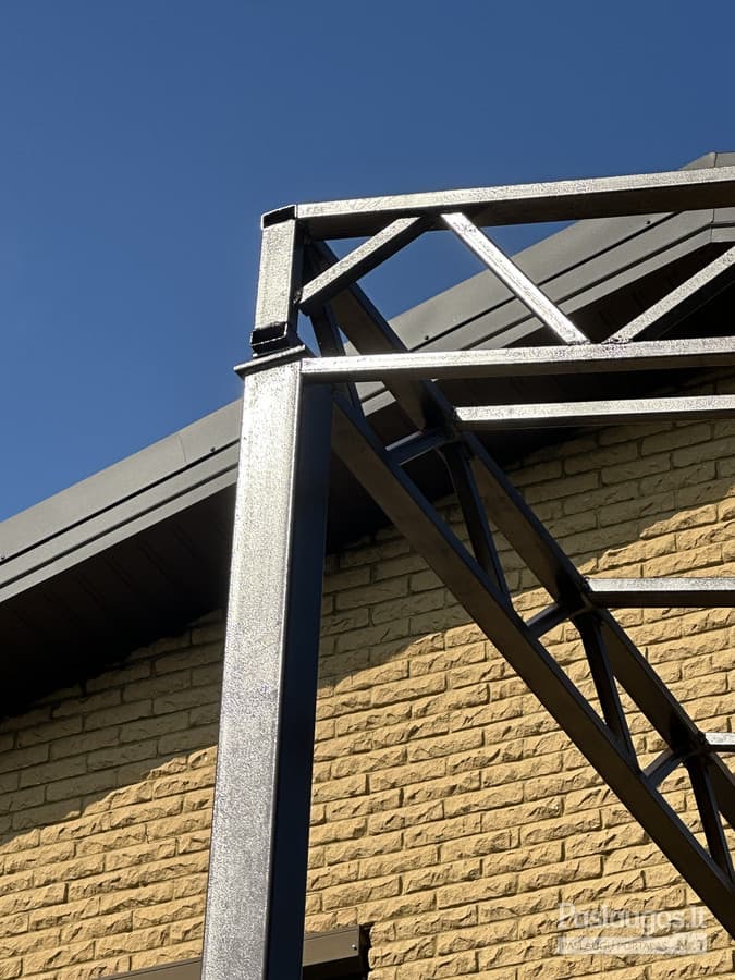 Plieno karkaso pergola – kampinis mazgas prieš mūrinio namo fone